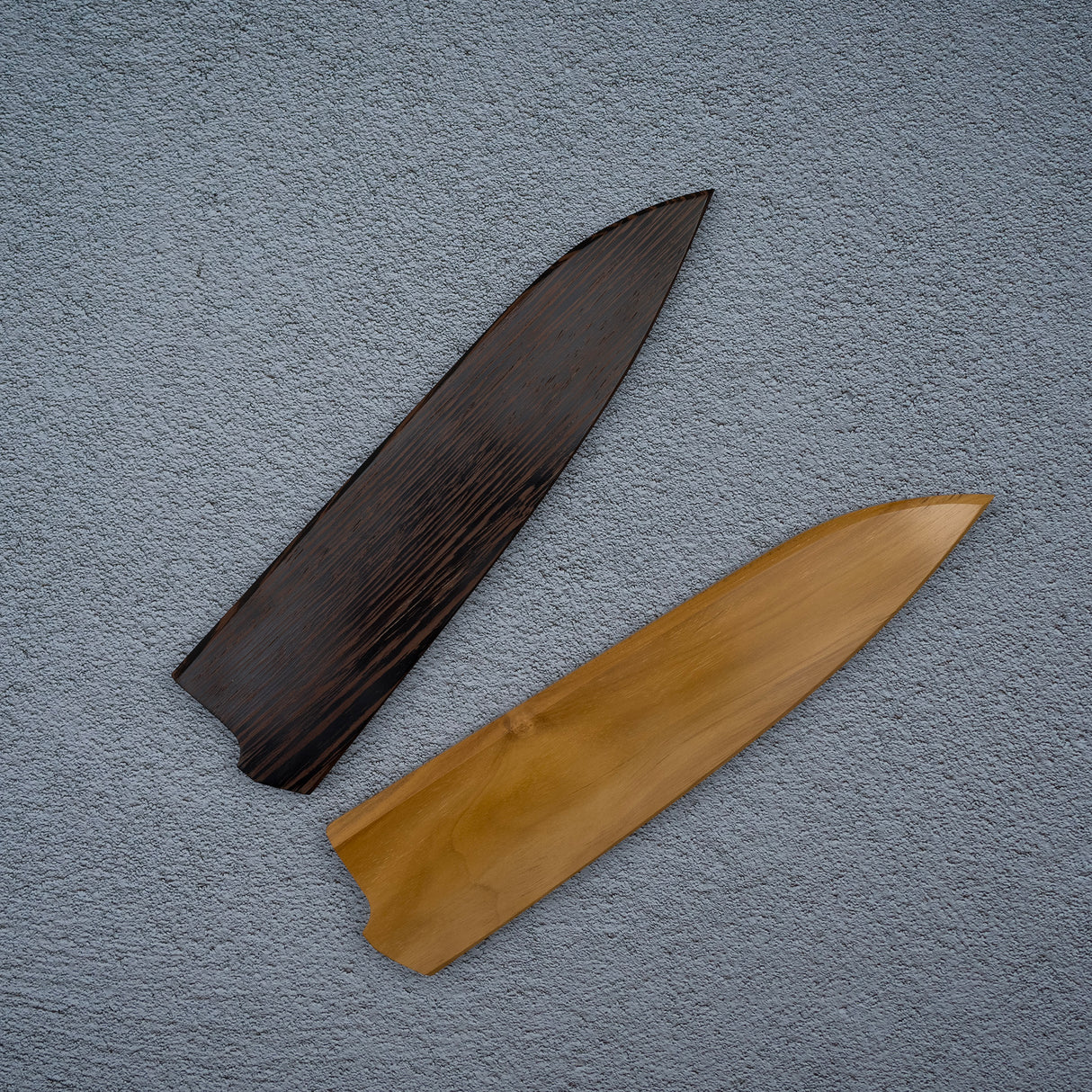 Two parts of a wooden knife saya laid on a grey surface, one dark brown and one light brown, possibly made of different types of wood such as Wenge and Teak.