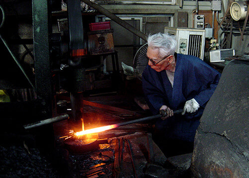 Keijiro Doi at the spring hammer in the Doi forge, drawing out a glowing billet with tongs.